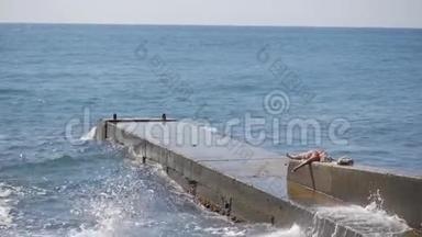 大浪在混凝土防波堤上破碎。 一个穿泳衣的胖女人躺在防波堤上晒黑。